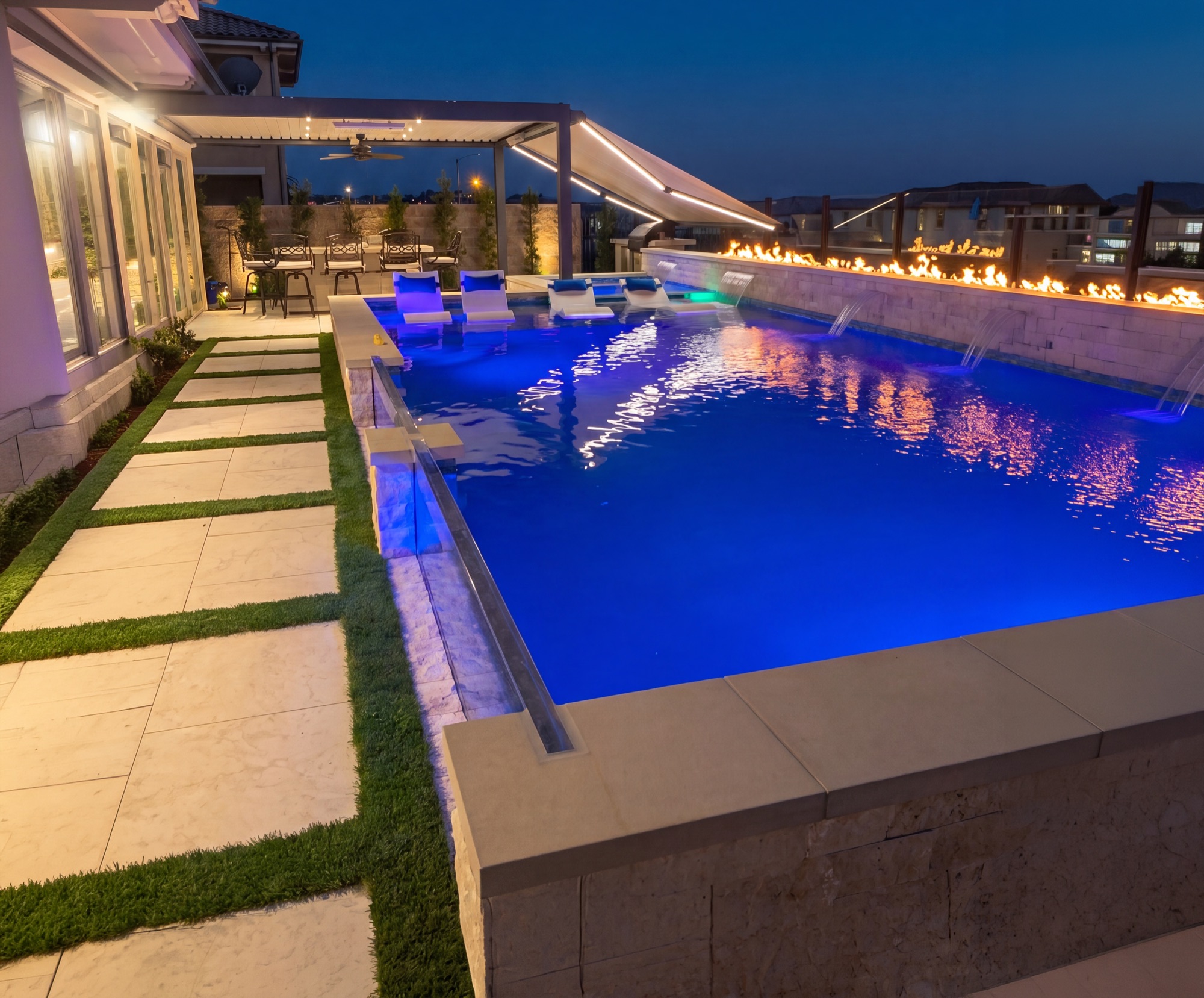 Pool with in-water loungers, fire feature and paver walkway at night in Baker Ranch, CA
