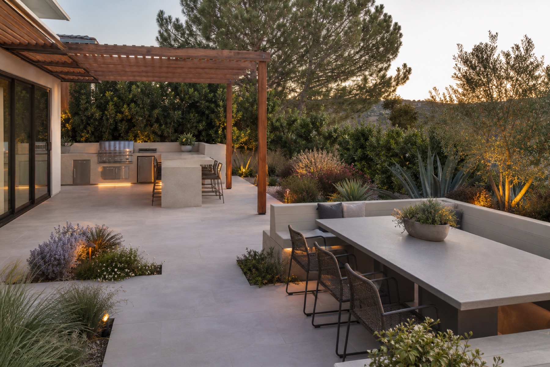 Modern backyard by Pacific Landscape Construction with pergola, outdoor kitchen, built-in dining seating, and drought-tolerant planting at dusk